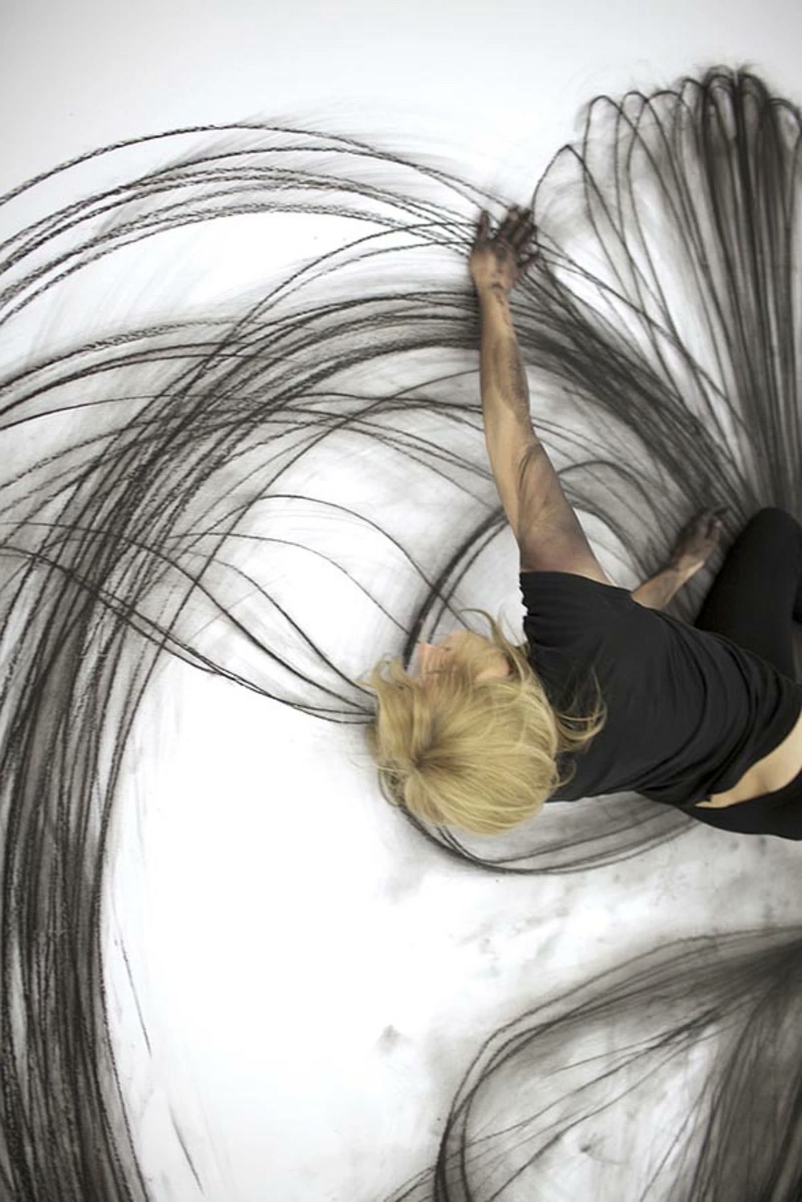 An artist sweeping long charcoal arcs across a white floor with her whole body